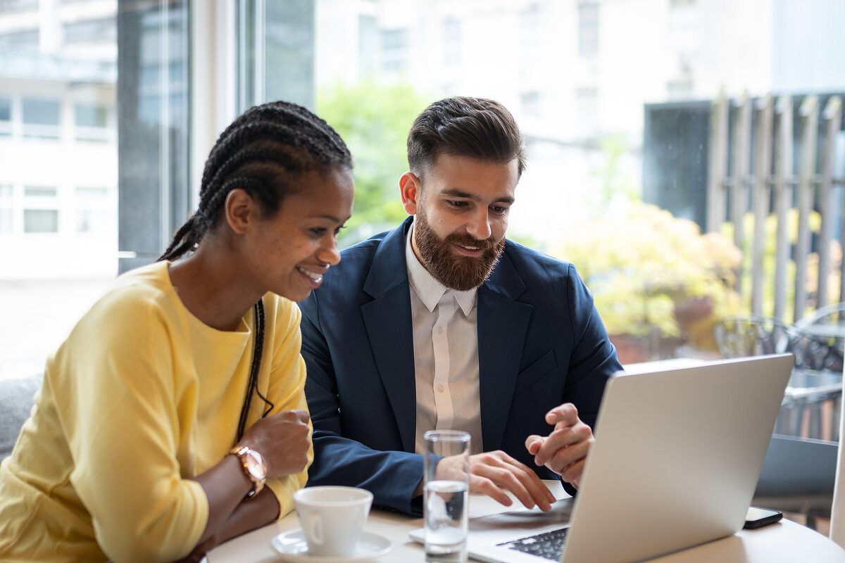 Two colleagues smile while reviewing information on a laptop in a bright café.