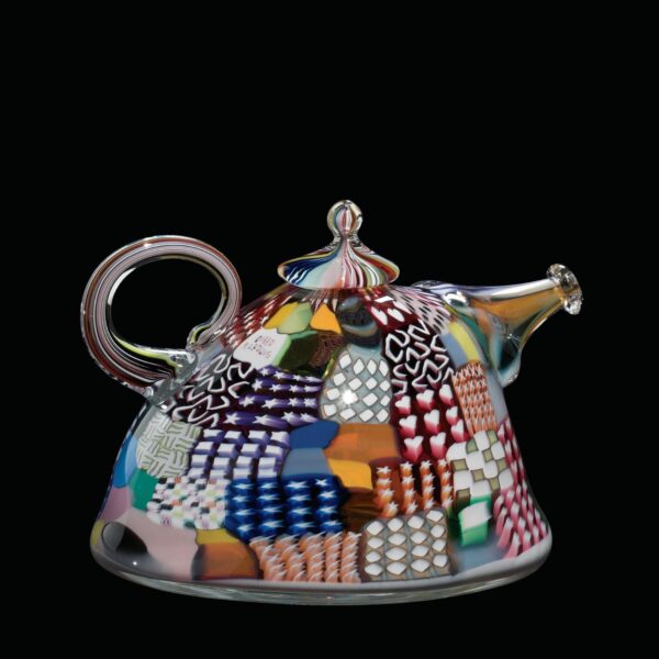 Glass teapot from 1980 decorated with colorful geometric patterns resembling quilt patchwork, against a black background.