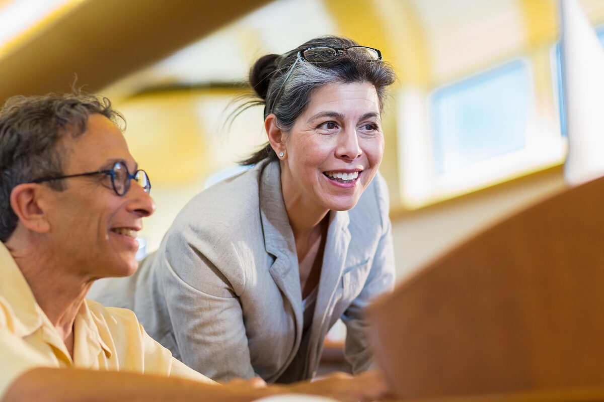Two colleagues smiling and collaborating in a bright, modern workspace, with one leaning forward to engage in discussion.