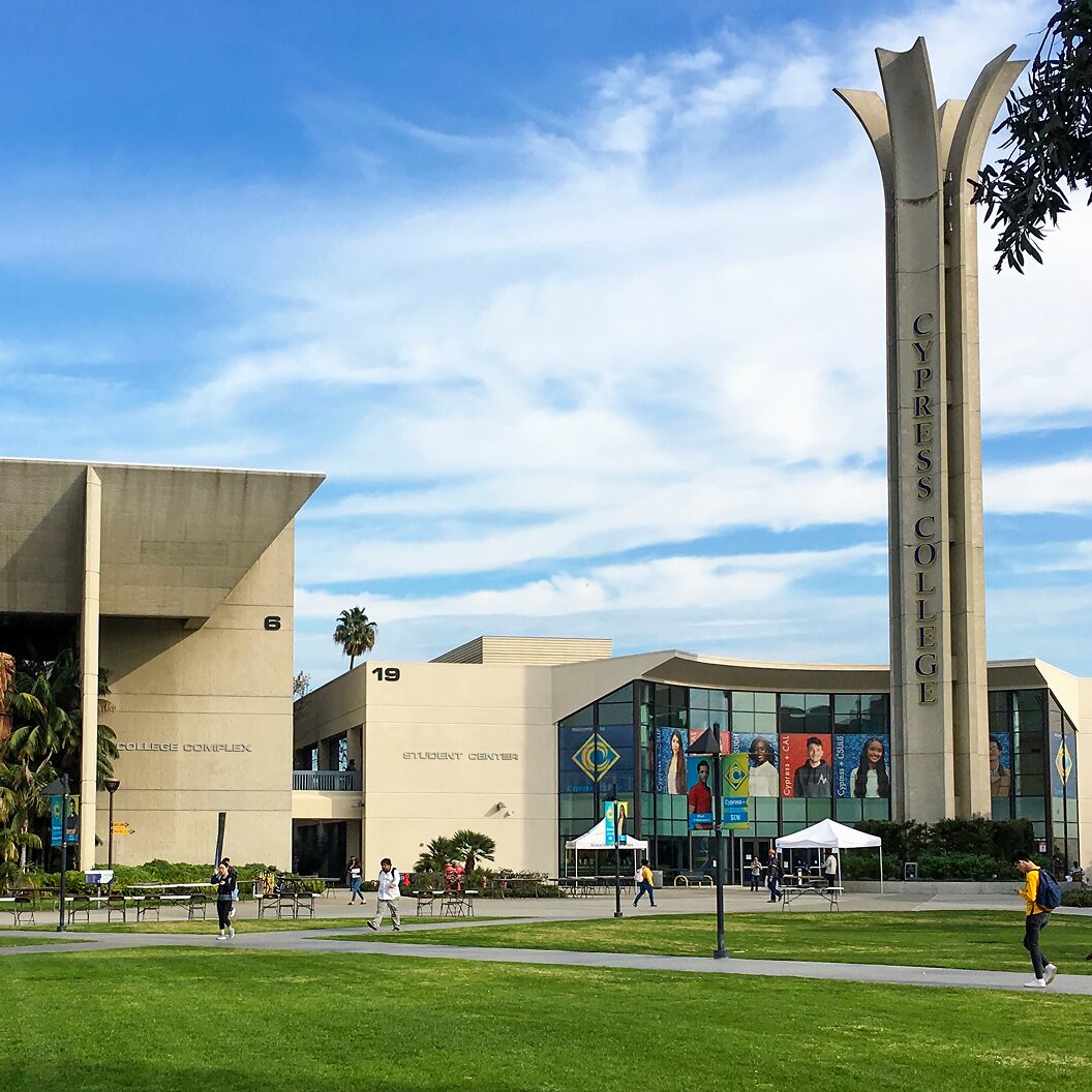 Exterior view of Cypress College campus with students walking across the lawn in front of modern buildings under a blue sky.
