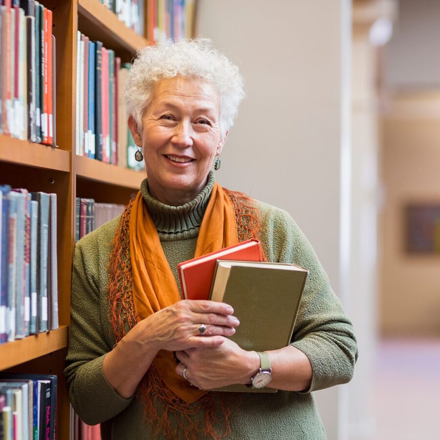 Smiling person standing in a library aisle, holding books against their chest.