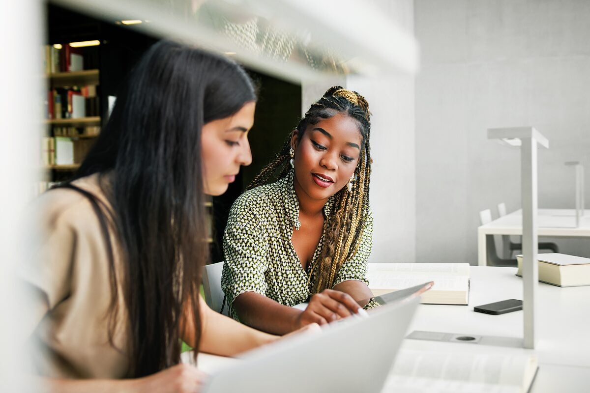 Two women collaborate at a library table, discussing content on a laptop.