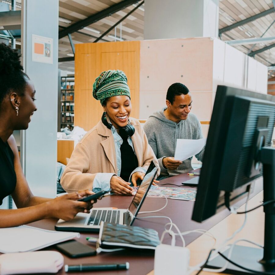 A group of three people work together around computers in a shared workspace, smiling and exchanging ideas.