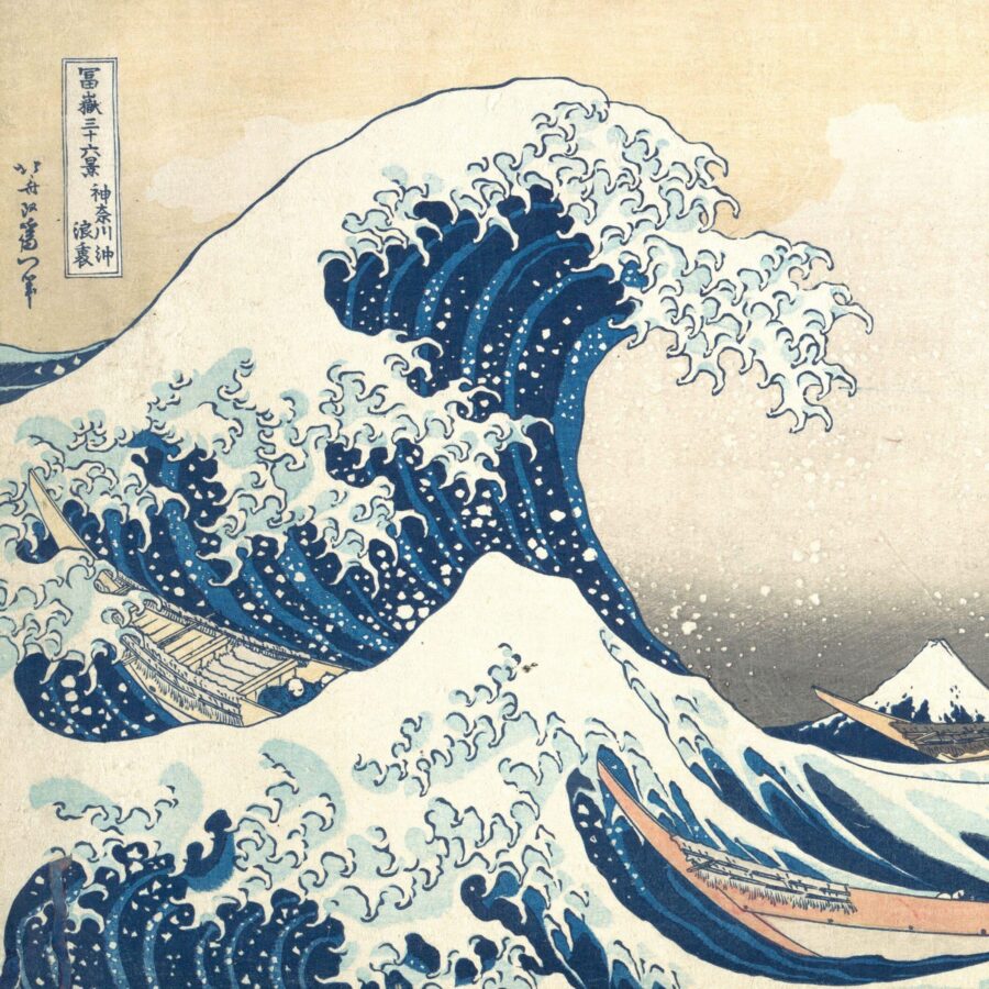 Japanese woodblock print showing a towering blue wave curling over small boats with rowers, with Mount Fuji visible in the distance beneath a pale sky.