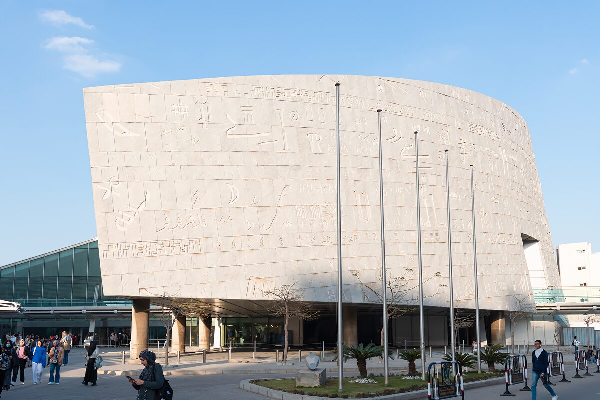 Exterior of a modern cultural building with stone walls inscribed with writing, people walking nearby on a sunny day.