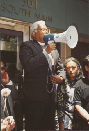 Color photo of a man in a clerical collar speaking through a blue megaphone outside a building marked “301 South Main,” with people listening.