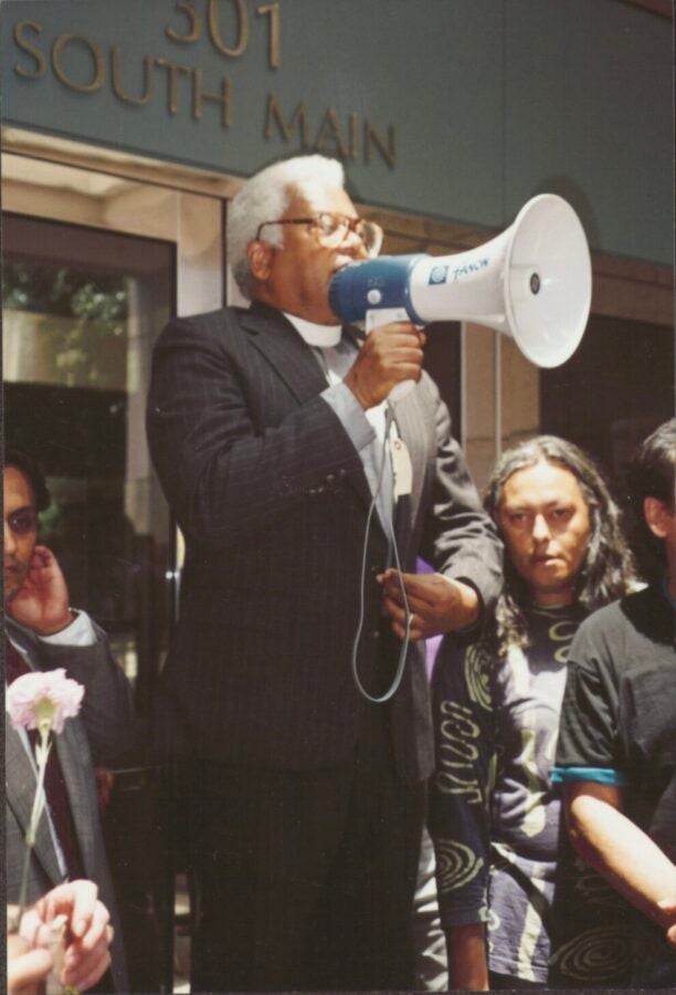 Color photo of a man in a clerical collar speaking through a blue megaphone outside a building marked “301 South Main,” with people listening.