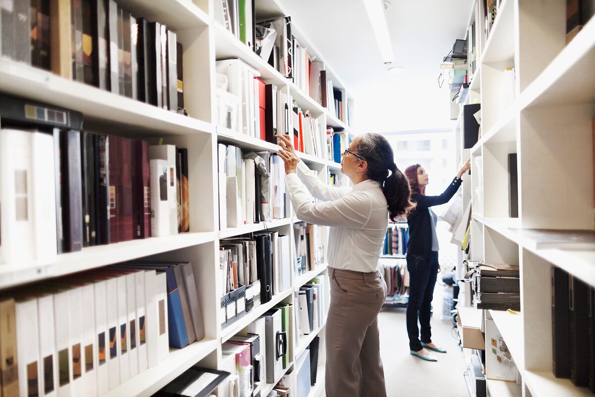 Two people browse library shelves, selecting books and research materials.