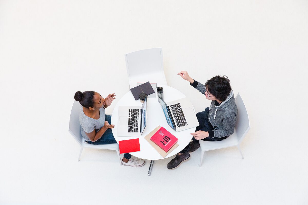 Overhead view of two students sitting at a round white table with laptops, notebooks, and coffee cups, engaged in discussion and collaboration.