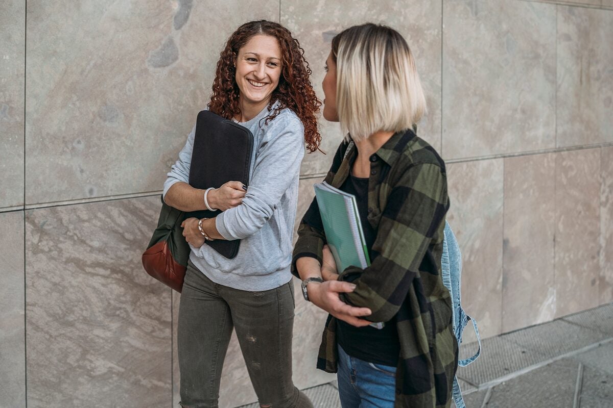 Two college students walking outside a campus building, smiling and talking while carrying notebooks and backpacks.