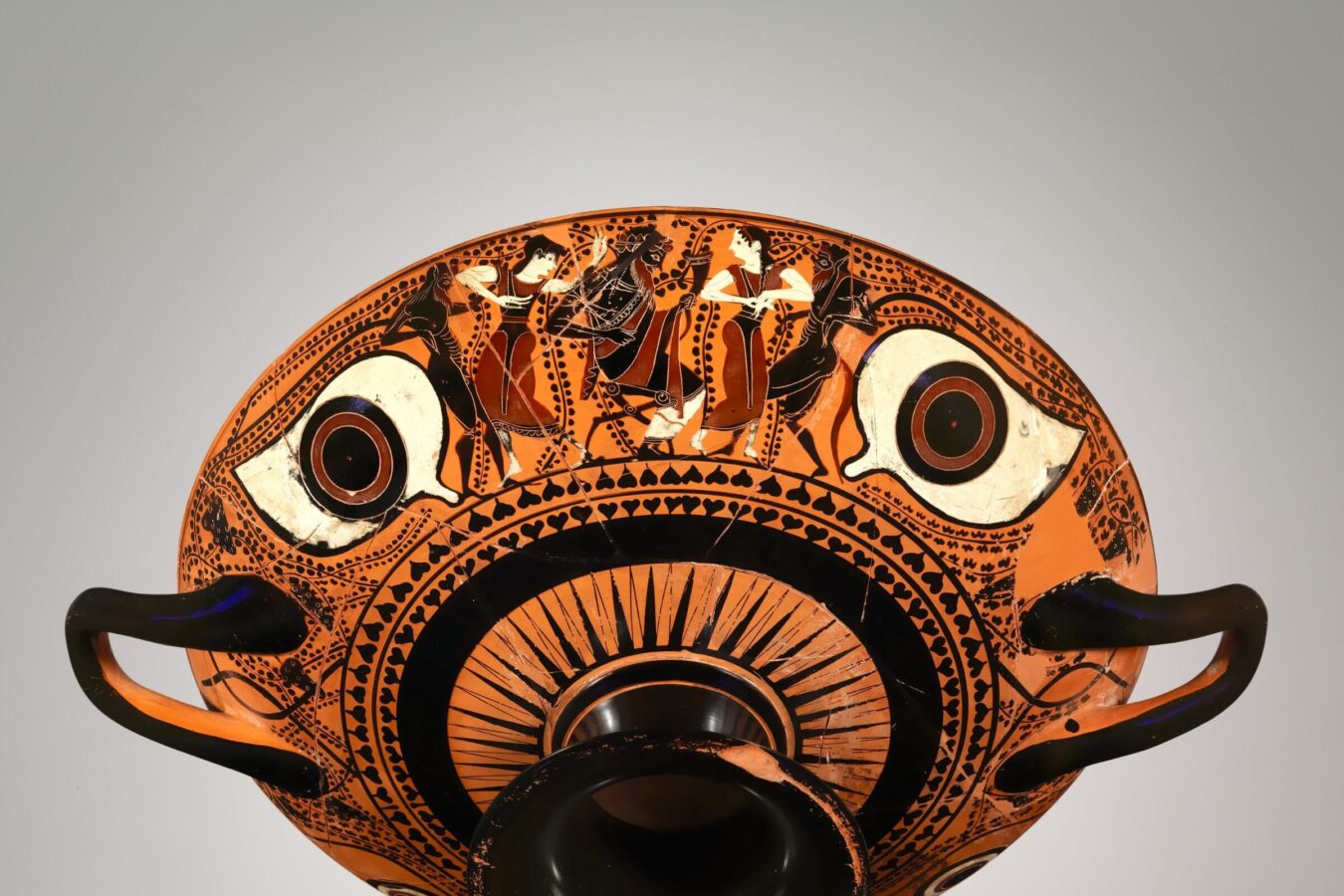 Ancient Greek terracotta kylix (drinking cup) painted in black and red with large stylized eyes and a central band depicting human figures in motion, bordered by geometric patterns.