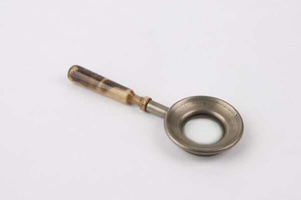 A vintage magnifying glass with a small circular lens set in a shallow metal dish and a short wooden handle. The handle has a smooth, polished finish with visible grain, and the metal dish is slightly tarnished, suggesting age and use. The object rests on a plain white background.