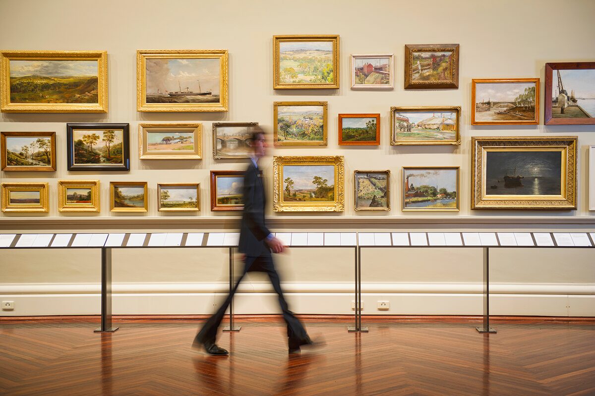 A person walks through an art gallery lined with framed paintings displayed on cream-colored walls.