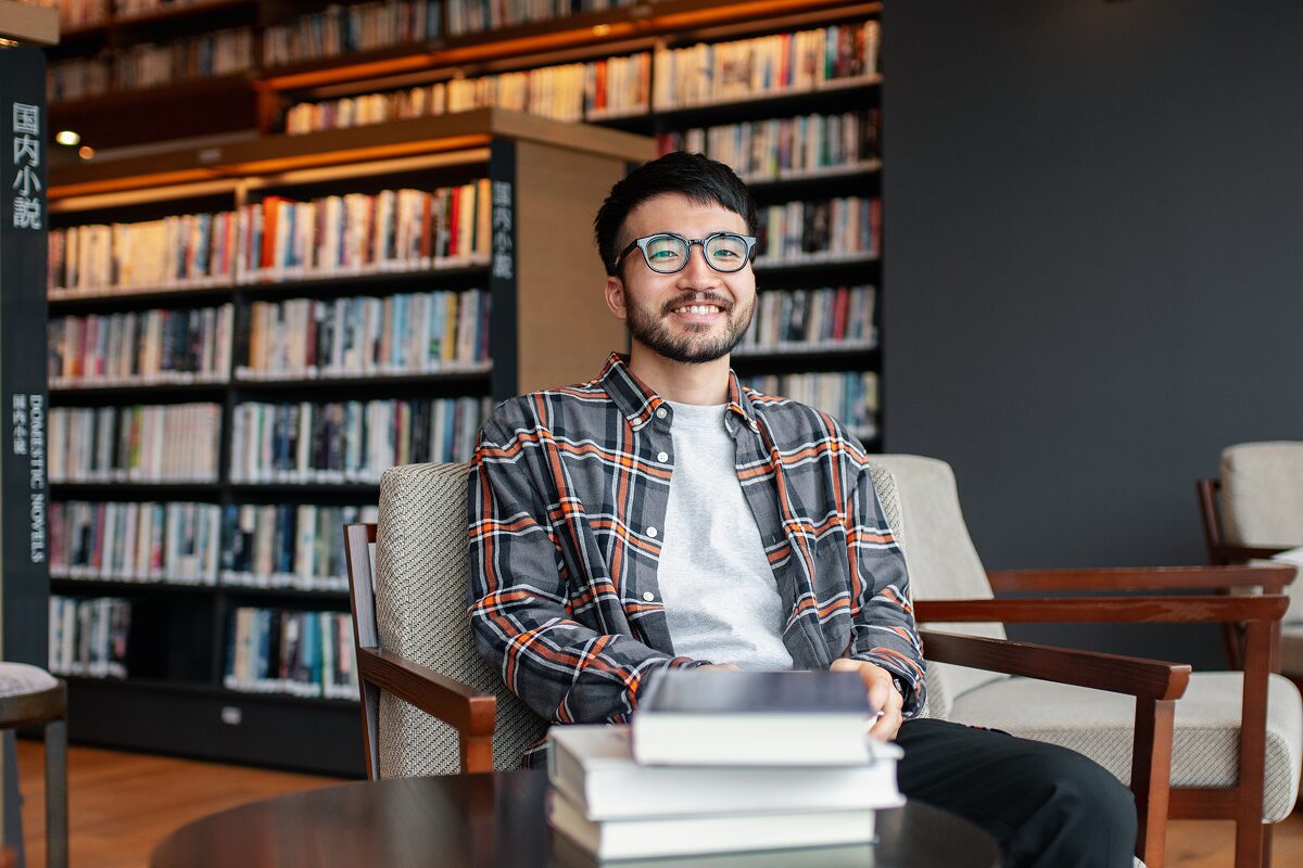 A smiling person sits in a library with stacked books on the table and full bookshelves in the background.
