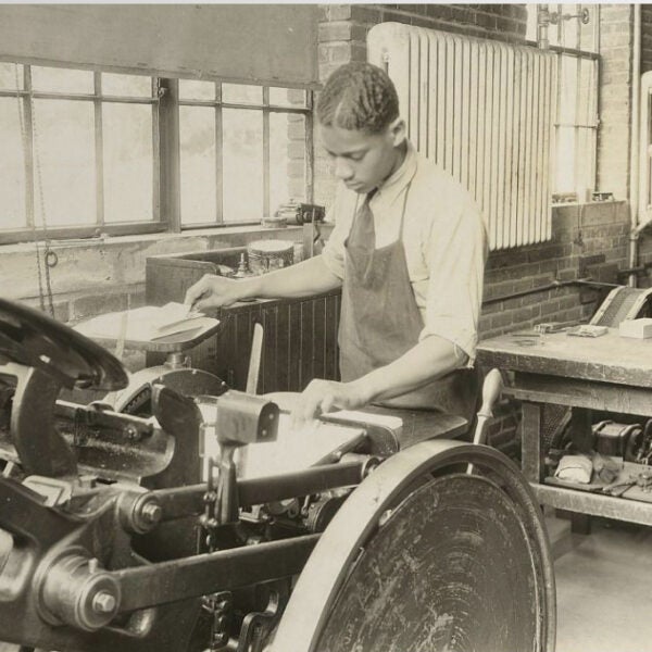 Black-and-white photograph of a man operating a printing press in a workshop. He wears a collared shirt and apron, feeding paper into the press beside work tables and machinery.