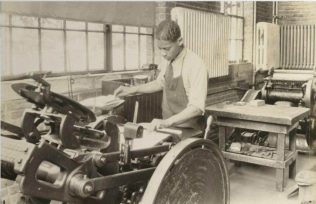 Black-and-white photograph of a man operating a printing press in a workshop. He wears a collared shirt and apron, feeding paper into the press beside work tables and machinery.