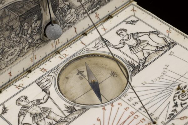 An intricately carved ivory sundial and compass in the form of a folding book. The open surface features engraved figures of men in period dress holding banners, with a central compass needle pointing north. Fine calibration lines and Roman numerals mark the sundial’s face, while a plumb line and detailed scene of workers appear on the raised lid.