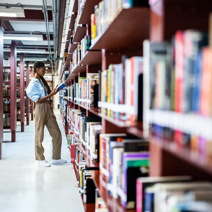 Person standing in a library aisle, selecting a book from a tall shelf.