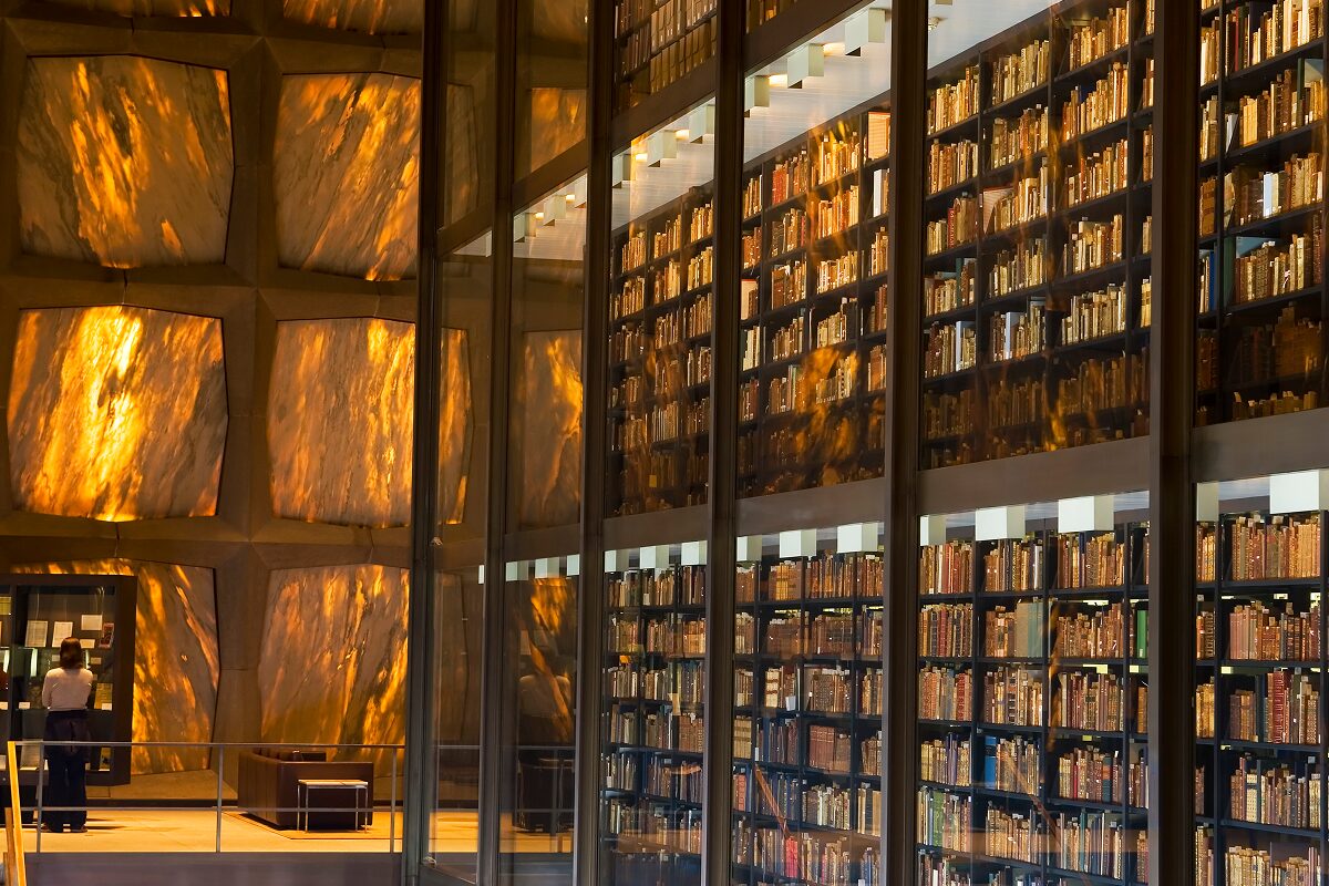 Interior view of a modern library with tall glass walls and multiple stories of bookshelves illuminated by warm light, reflecting on the surrounding stone walls.