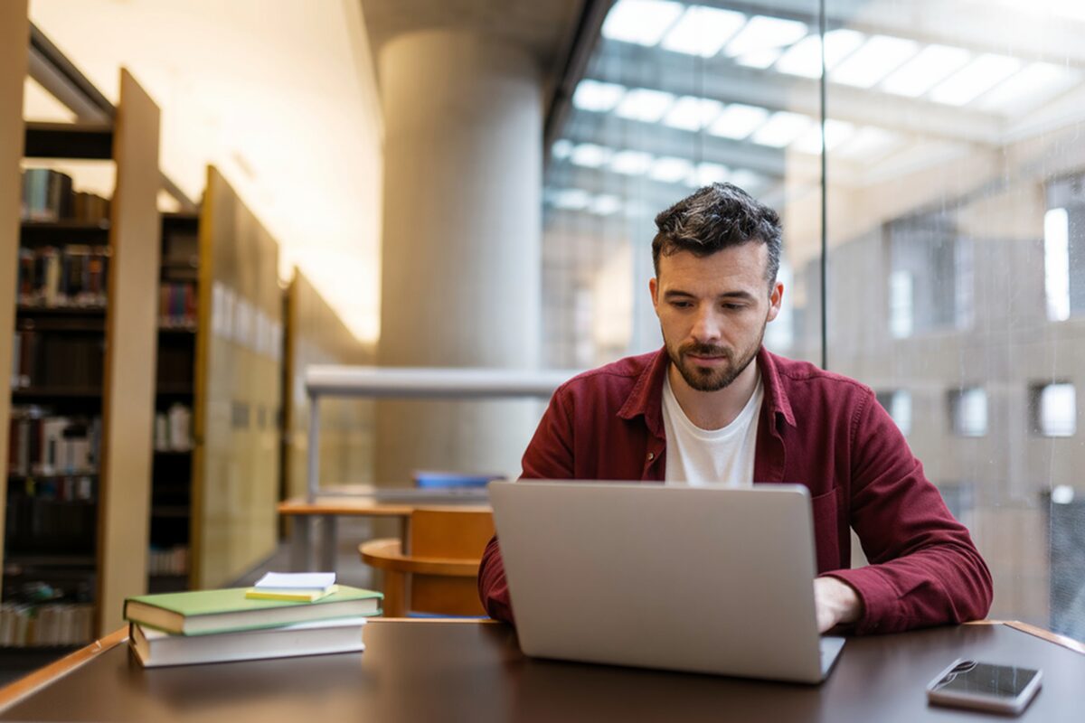 Person working on a laptop at a library table with books and natural light coming through large windows.