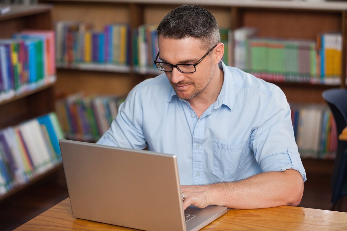 Person working on a laptop at a wooden desk in a library with colorful books on the shelves behind them.