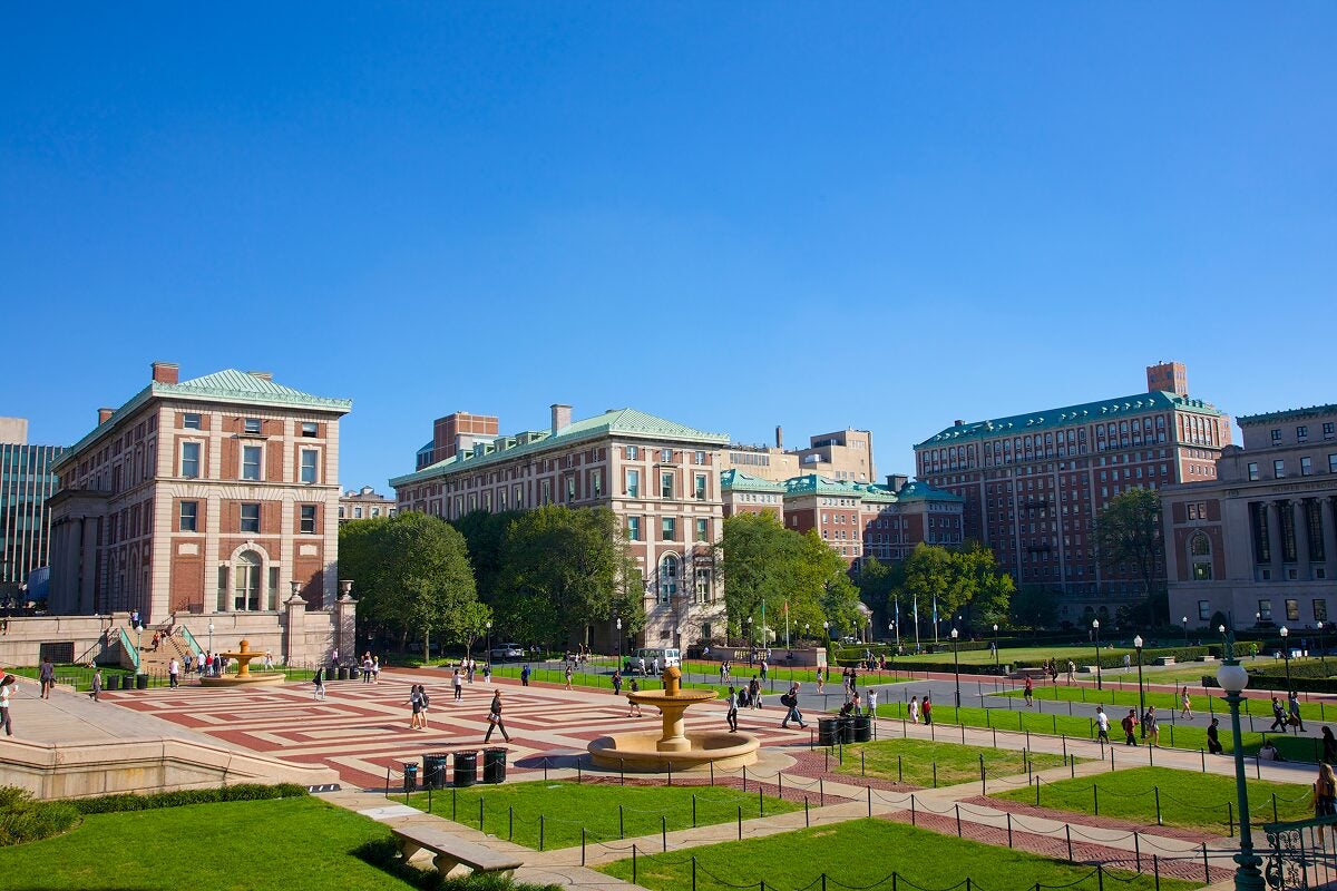 Wide view of a large university campus with students walking across a plaza bordered by classic red-brick buildings and green lawns under a clear blue sky.