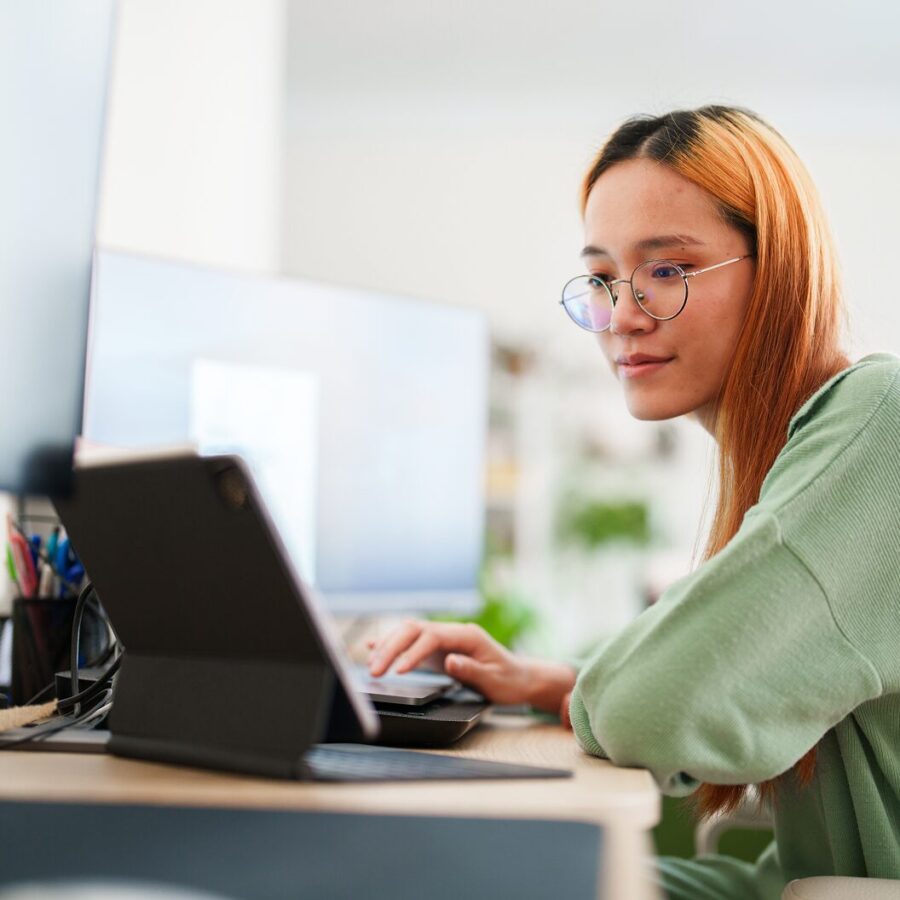 Person wearing glasses using a tablet and laptop at a desk in a bright home workspace.