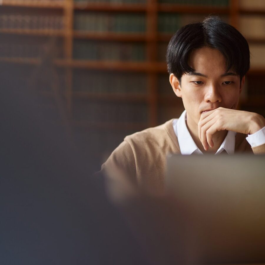 Person studying in a library, focused on a laptop with shelves of books in the background.