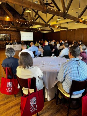 Attendees seated at round tables listen to a speaker at a podium in a large, rustic room with string lights and exposed beams. A presentation slide on the screen reads, “What do you see as the most significant challenge facing ebook publishing today?” Participants appear engaged, with red JSTOR 30th anniversary tote bags visible around the room.