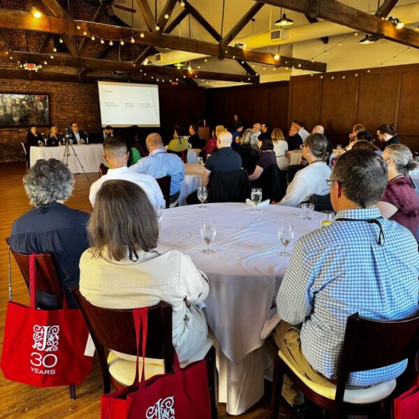 Attendees seated at round tables listen to a speaker at a podium in a large, rustic room with string lights and exposed beams. A presentation slide on the screen reads, “What do you see as the most significant challenge facing ebook publishing today?” Participants appear engaged, with red JSTOR 30th anniversary tote bags visible around the room.