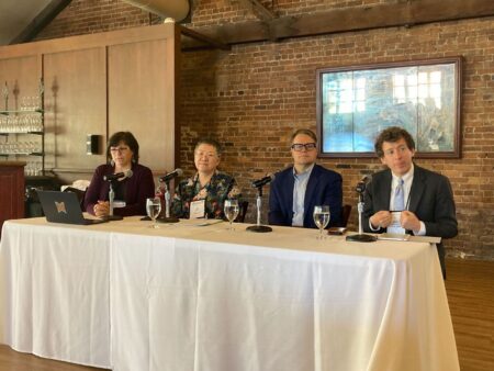 Four panelists sit behind a white table, each with a microphone and water glass. The panel includes two women and two men, seated against a brick wall with a framed picture in the background. The atmosphere is professional and collegial, suggesting an academic or publishing discussion.