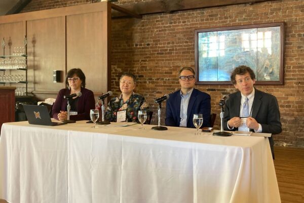 Four panelists sit behind a white table, each with a microphone and water glass. The panel includes two women and two men, seated against a brick wall with a framed picture in the background. The atmosphere is professional and collegial, suggesting an academic or publishing discussion.