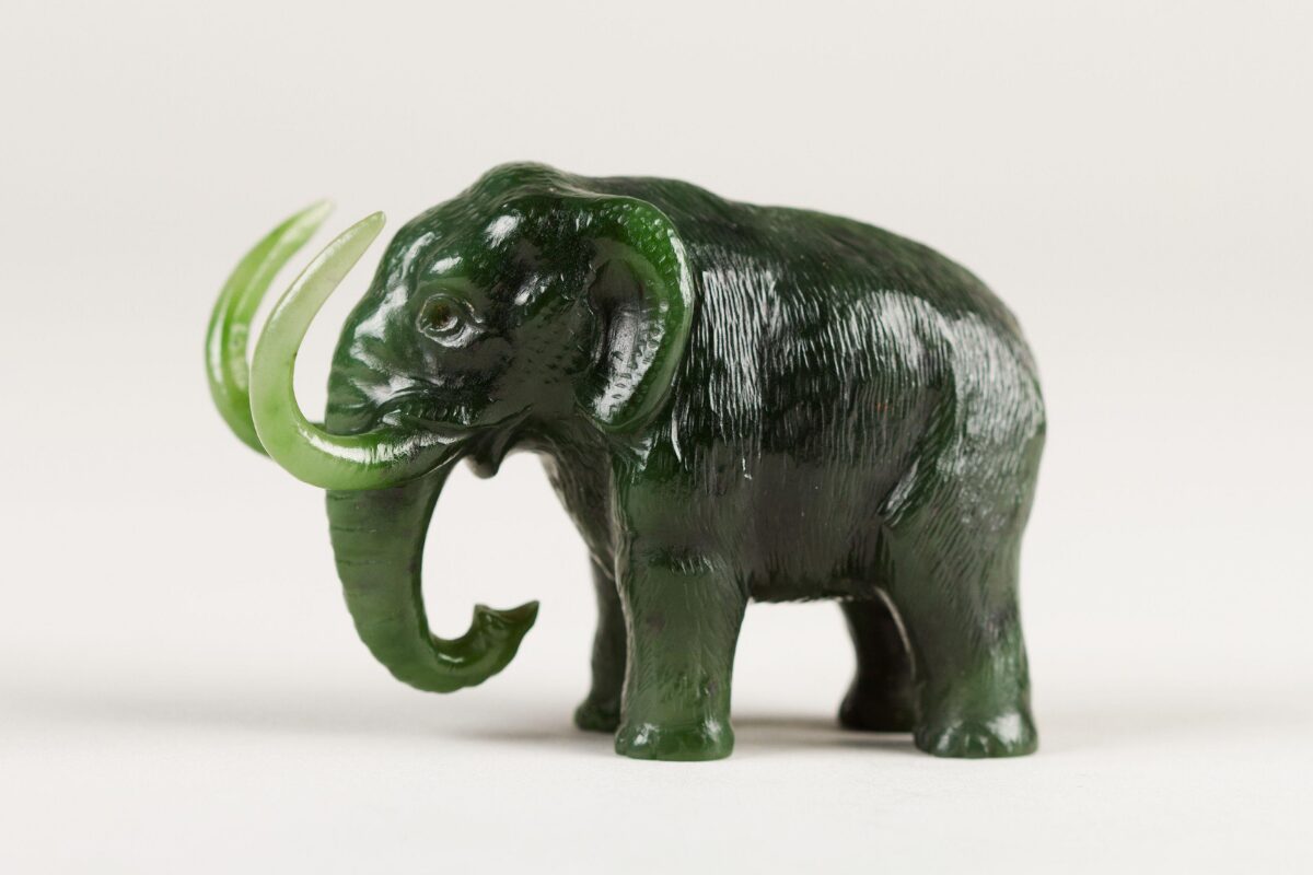 A small dark green carved figurine of an elephant with lighter green tusks and a curled trunk, shown against a plain light background.