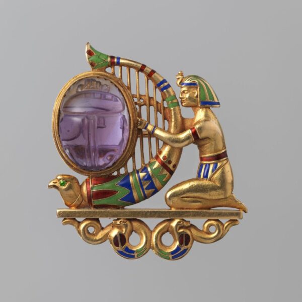 A gold Egyptian Revival brooch featuring a kneeling figure beside a harp-like frame holding a purple carved gemstone, with colorful enamel details.