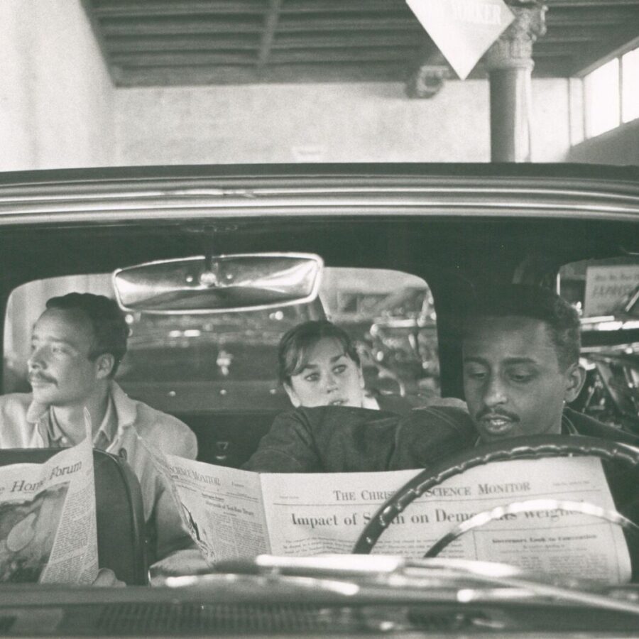 Black-and-white photo of three people sitting in a parked car, each holding a newspaper; the two men in the front seat read, while the woman in the back looks forward between them.