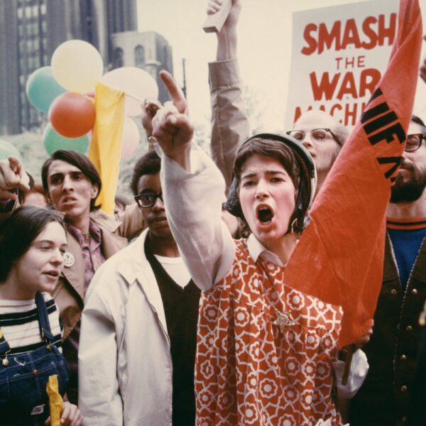 A young protester in a patterned red tunic raises her fist and shouts amid a crowd of demonstrators holding signs and balloons during an anti–war march.