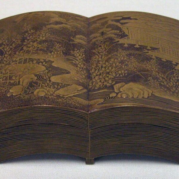 A brown lacquer box shaped like an open book, decorated with intricate gold landscape designs of plants, rocks, and distant buildings.