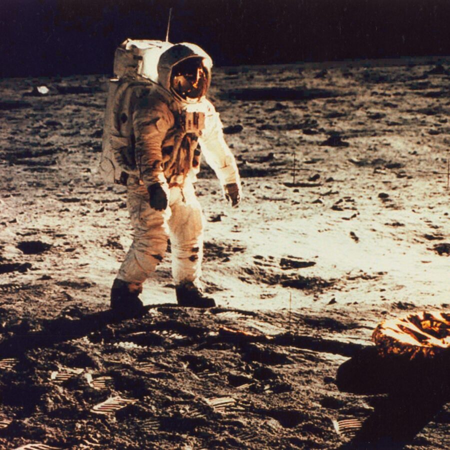 Astronaut in a white spacesuit standing on the moon’s surface near a lander footpad, with footprints and lunar soil illuminated against the dark sky.
