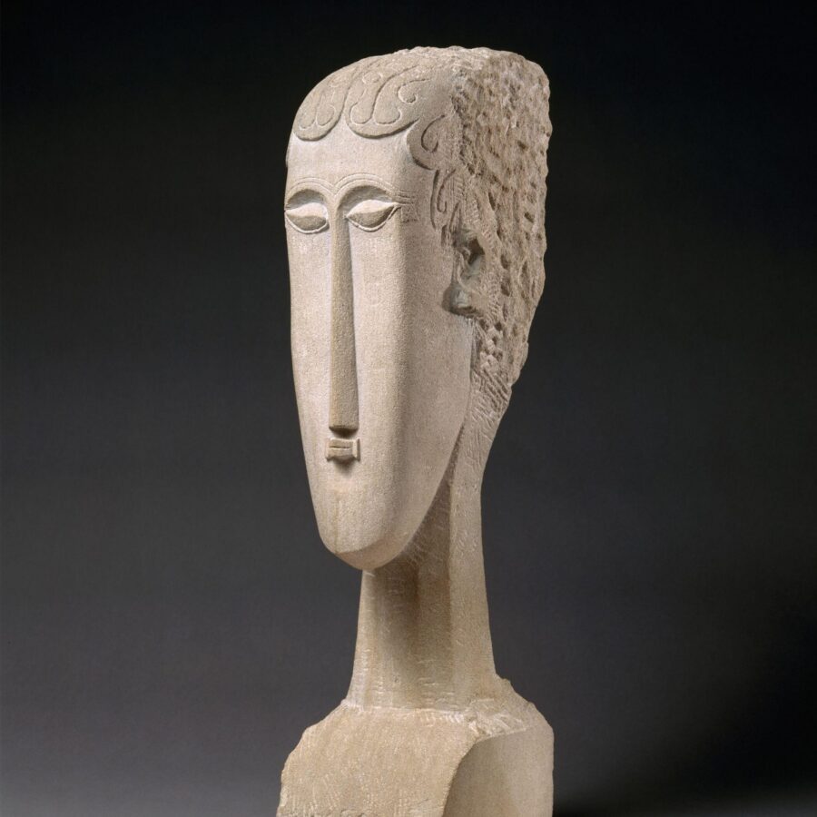A carved stone bust with an elongated face, stylized features, and a tall narrow nose, reminiscent of Amedeo Modigliani’s sculptural style.