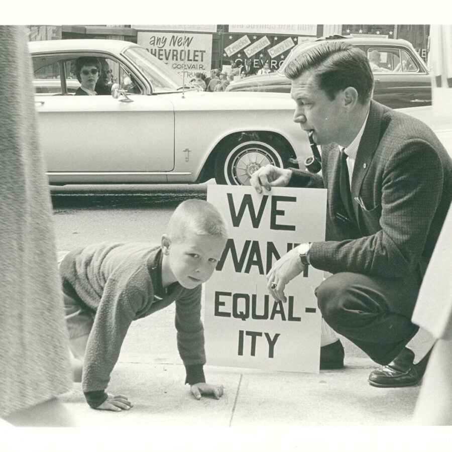 A man kneels beside a young boy holding a protest sign reading “We want equality,” while a woman watches from a nearby car.