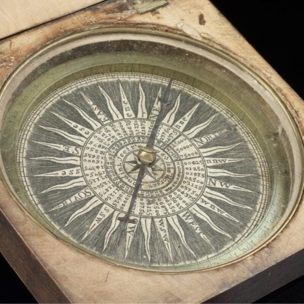 A 17th-century magnetic compass housed in a wooden case with brass hinges. The compass features a detailed wind rose with cardinal directions and degree markings engraved on a circular plate. The needle points north, and the ornate design includes concentric rings of numbers and letters used for navigation.