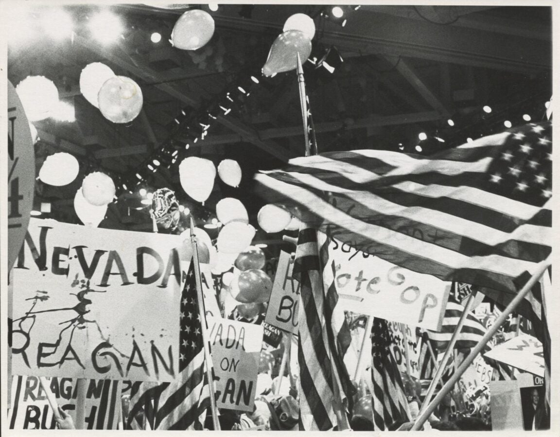 A crowded political convention filled with American flags, balloons, and handmade signs supporting Reagan and Bush. Delegates hold state signs such as “Nevada,” while balloons float near the ceiling amid bright overhead lights.