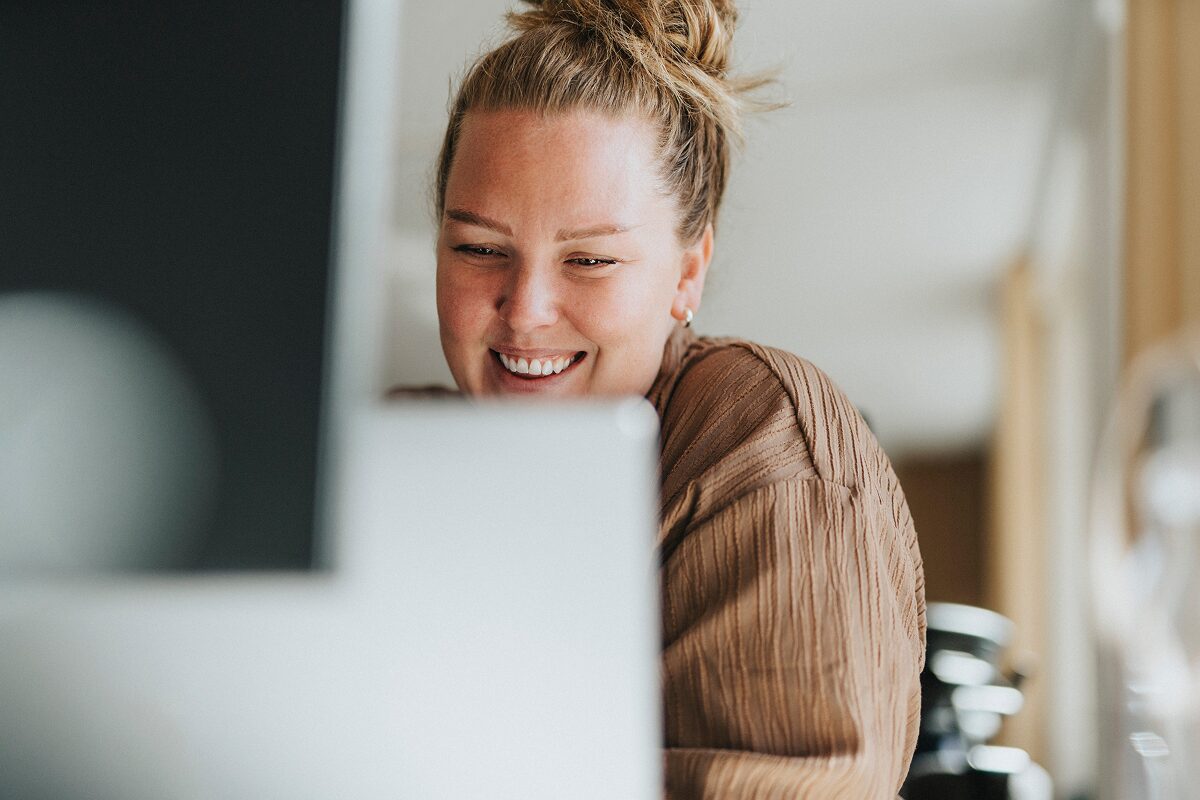 Smiling woman with hair in a bun working at a laptop in a bright, modern indoor setting.