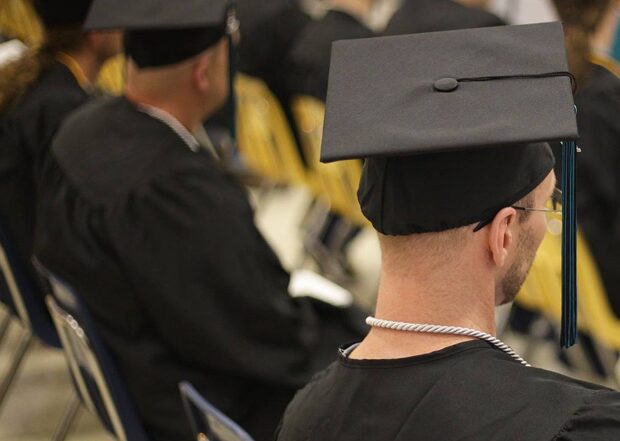 A room of people in graduation gowns and caps sitting with their backs to the camera, facing a speaker.