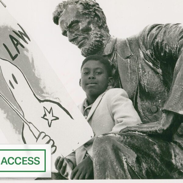 Black-and-white photo of a young boy sitting beside a large Lincoln statue, holding a protest sign depicting a hooded figure labeled “The Law,” with an “Open Access” badge.
