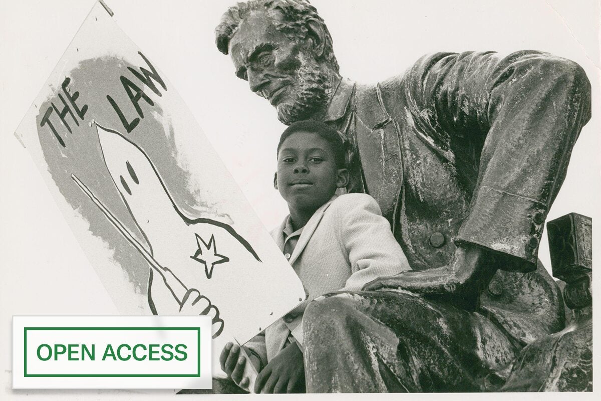 Black-and-white photo of a young boy sitting beside a large Lincoln statue, holding a protest sign depicting a hooded figure labeled “The Law,” with an “Open Access” badge.