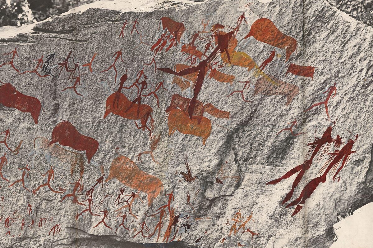 Rock surface covered with vivid red and orange ancient rock paintings depicting human figures running, hunting, and riding animals among stylized cattle and wildlife.
