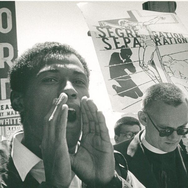 A man shouts during a civil rights demonstration, with a large protest sign about segregation visible behind him.