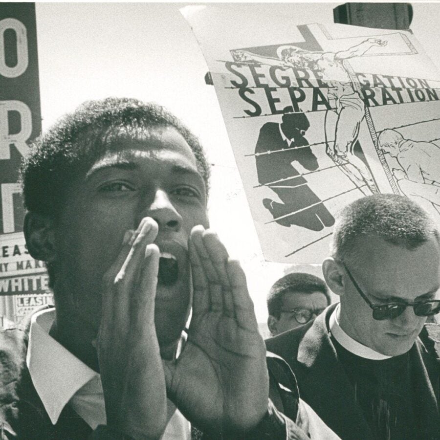 A man shouts during a civil rights demonstration, with a large protest sign about segregation visible behind him.