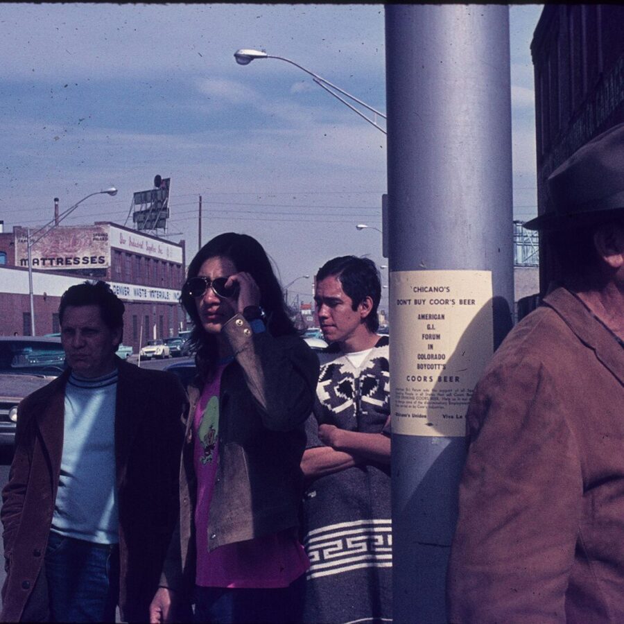 A group of young people stand on a city sidewalk near a pole with a flyer urging Chicanos to boycott Coors beer; industrial buildings and cars line the street in the background.